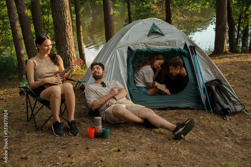 Shot of joyful company of friends with guitar camping in summer forest.