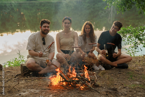 Shot of friends hikers with beer and guitar frying sausages near river in forest trip.