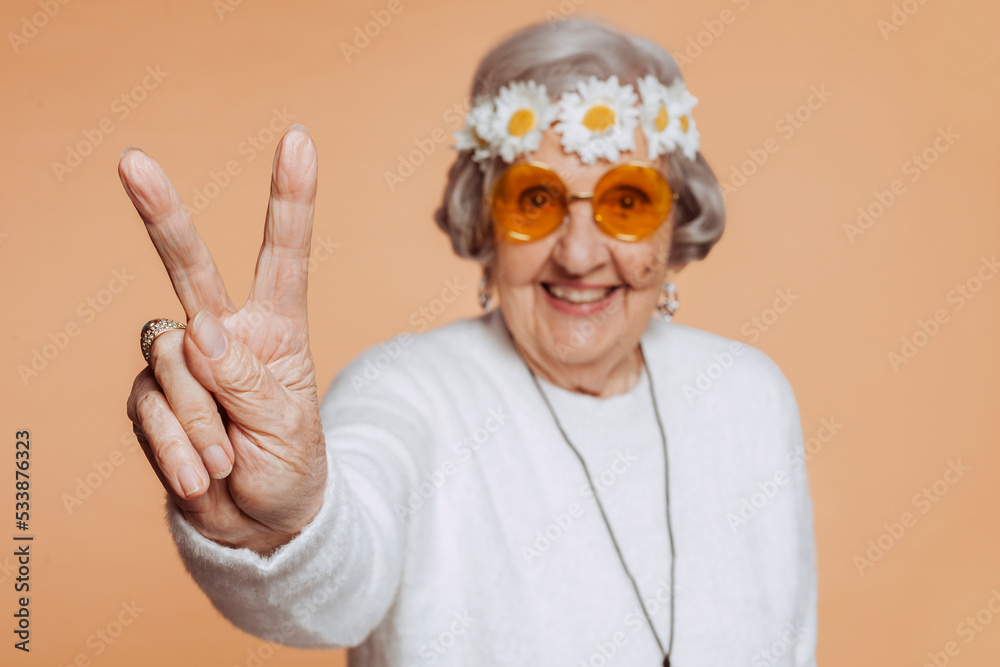 Close-up portrait of senior old smiling grandmother does peace victory sign with fingers. Hippie woman with wreath on her head. Close-up portrait of senior old smiling grandmother does peace victory sign with fingers. Hippie woman with wreath on her head.