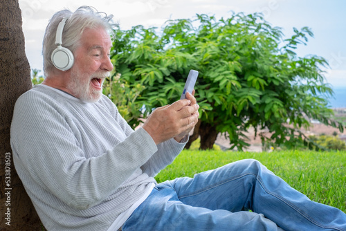 Happy senior man wearing headphones sitting on the meadow in public park having fun in video call by smartphone.