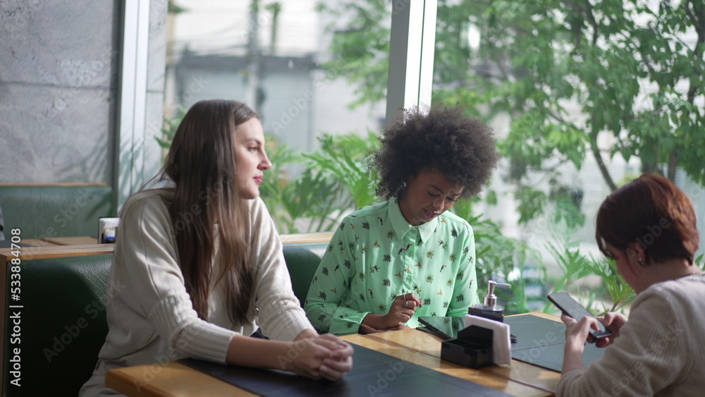 Candid group of friend seated at coffee shop. Diverse women at cafe restaurant. Three girlfriends at restaurant looking at gadgets Candid group of friend seated at coffee shop. Diverse women at cafe restaurant. Three girlfriends at restaurant looking at gadgets