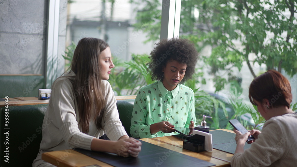 Candid group of friend seated at coffee shop. Diverse women at cafe restaurant. Three girlfriends at restaurant looking at gadgets Candid group of friend seated at coffee shop. Diverse women at cafe restaurant. Three girlfriends at restaurant looking at gadgets