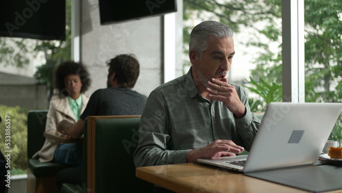 Middle aged man preparing to work adjusting shirt and organizing coat and items in front of laptop seated at coffee shop