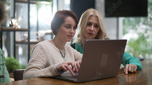 Female executive woman explaining job to young female employee in front of laptop at cafe. Female entrepreneur in conversation with staff