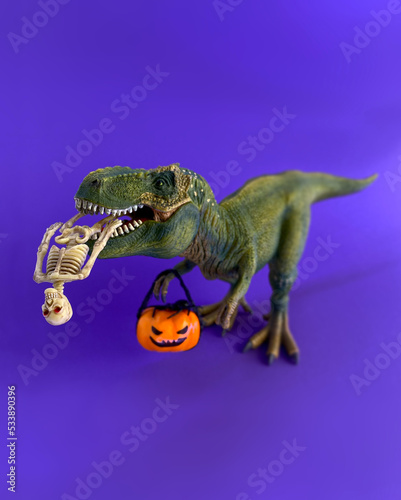 Happy Halloween - funny toy t-rex dinosaur with pumpkin jack-o-lantern and a human skeleton. Tyrannosaurus holding pumpkin, trick or treat. Purple background.