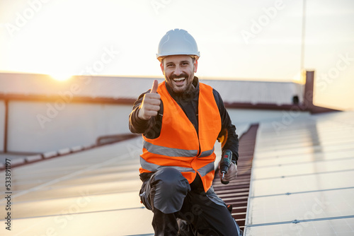 A happy worker with drill in his hands is giving thumbs up for using solar panels while holding a drill and smiling at the camera.
