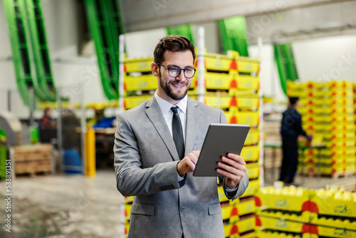 A business owner monitoring works in fruit production factory warehouse over the tablet and smiling at it.