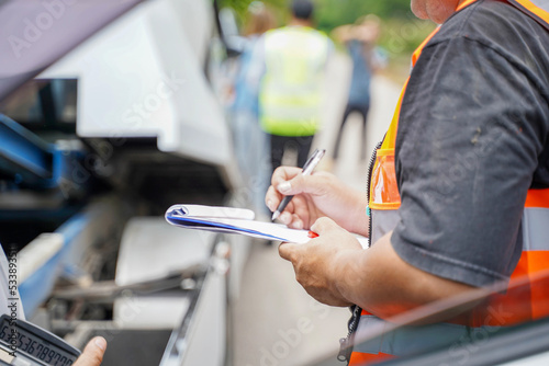Closeup and crop insurance agent writing on clipboard while examining car after accident claim being assessed and processed on blurred damaged car truck slides and group of people background.