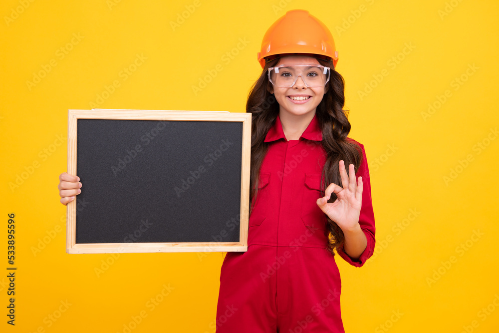 Child girl builder in hard hat helmet. Teenage girl worker hold blackboard isolated on yellow background. Kids renovation concept. Copy space, mock up. Child girl builder in hard hat helmet. Teenage girl worker hold blackboard isolated on yellow background. Kids renovation concept. Copy space, mock up.
