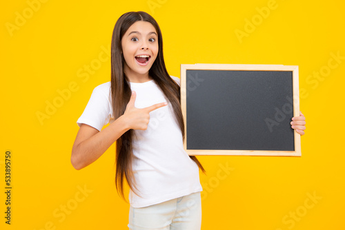Teenager younf school girl holding school empty blackboard isolated on yellow background. Portrait of a teen female student. Excited teenager, glad amazed and overjoyed emotions.