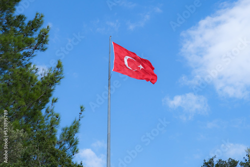 Flag of the Republic of Turkey on a high flagpole against a blue sky and clouds. The flag of Turkey flutters in the wind.