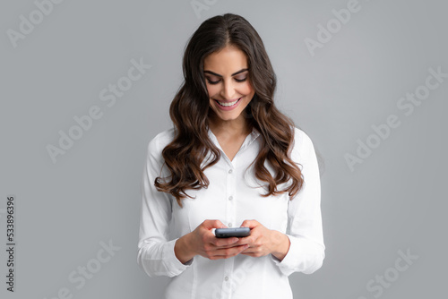 Smiling young brunette business woman girl in shirt isolated on gray background, studio portait. Beauty girl using mobile phone, typing sms message.