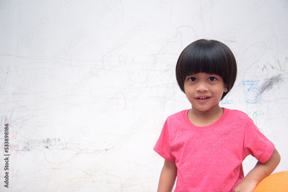 A smiling Asian youngster carrying an orange balloon while donning a pink outfit. On the house's white wall, he painted with several crayons. copy space. A smiling Asian youngster carrying an orange balloon while donning a pink outfit. On the house's white wall, he painted with several crayons. copy space.