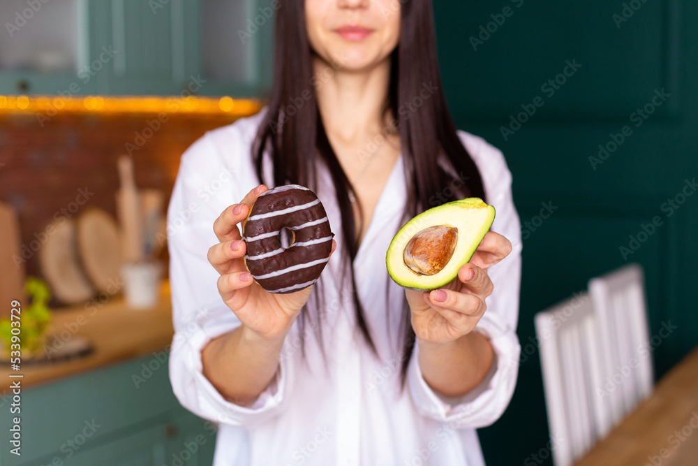 a young woman in a white shirt stands in the kitchen with a donut in one hand and an avocado in the other hand, makes a choice a young woman in a white shirt stands in the kitchen with a donut in one hand and an avocado in the other hand, makes a choice