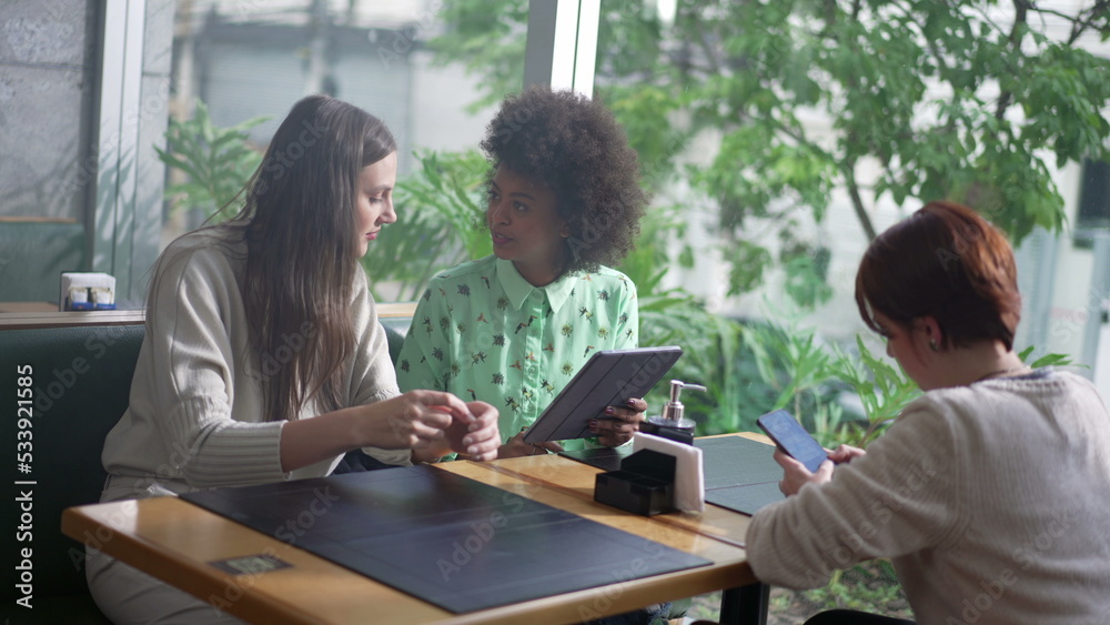 Woman showing tablet screen to female friend at coffee shop table. Three women interaction using modern technology devices Woman showing tablet screen to female friend at coffee shop table. Three women interaction using modern technology devices