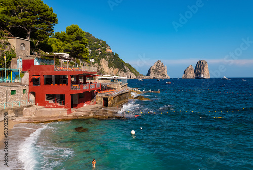 Fototapeta Naklejka Na Ścianę i Meble -  Capri summer panorama in “Marina piccola“, a small bathing spot on the coast of the italian island. Beach bars in a bay with 3 iconic landmark rocks “Faraglioni“ and big yachts in the background.