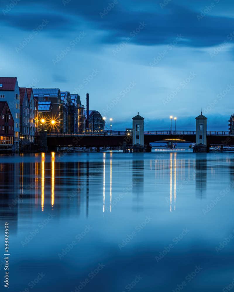 The harbor and city of Trondheim in Norway. Riverside with historic buildings and bridge over river with glowing street lanterns at blue hour. Reflection of night lights in smooth water surface.
