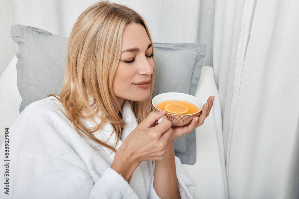 Pretty caucasian woman in white bathrobe drinking hot lemon tea enjoying spa procedures in wellness center Pretty caucasian woman in white bathrobe drinking hot lemon tea enjoying spa procedures in wellness center