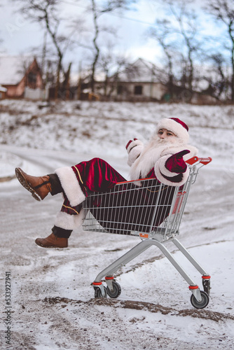a woman in Santa Claus costume in a snowy forest in winter with a wheelbarrow from a supermarket trolley 