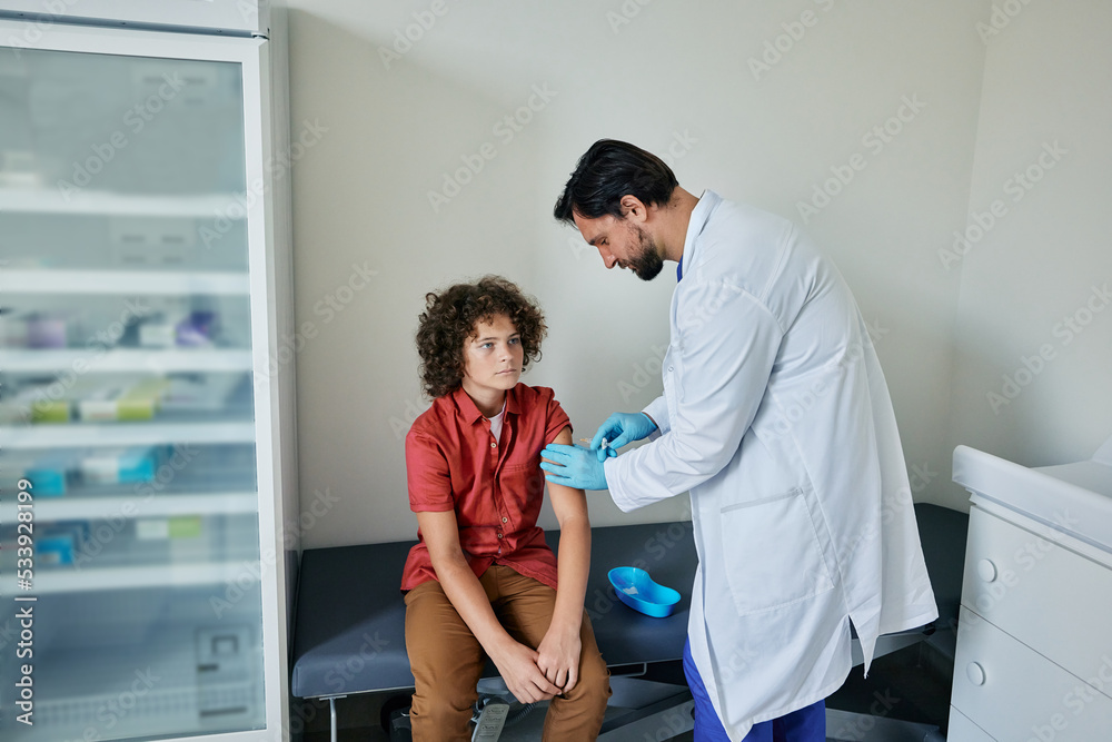 Routine vaccinations for child. Curly haired boy while vaccinated by male pediatrician at medical clinic Routine vaccinations for child. Curly haired boy while vaccinated by male pediatrician at medical clinic
