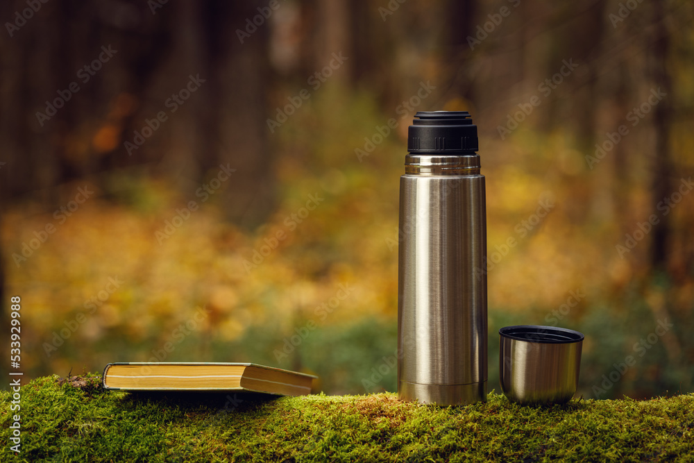 Steel thermos with delicious hot tea and a book on an old mossy