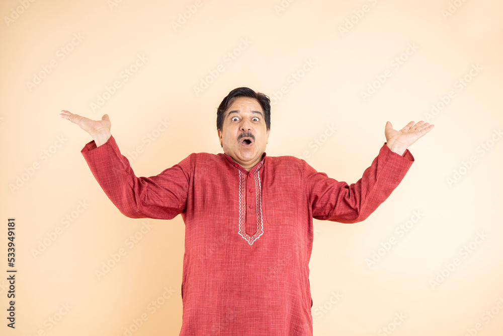 Amazed or shocked mature indian man with open mouth and astonished expression and open arms isolated on plane studio background. He is wearing traditional red kurta,