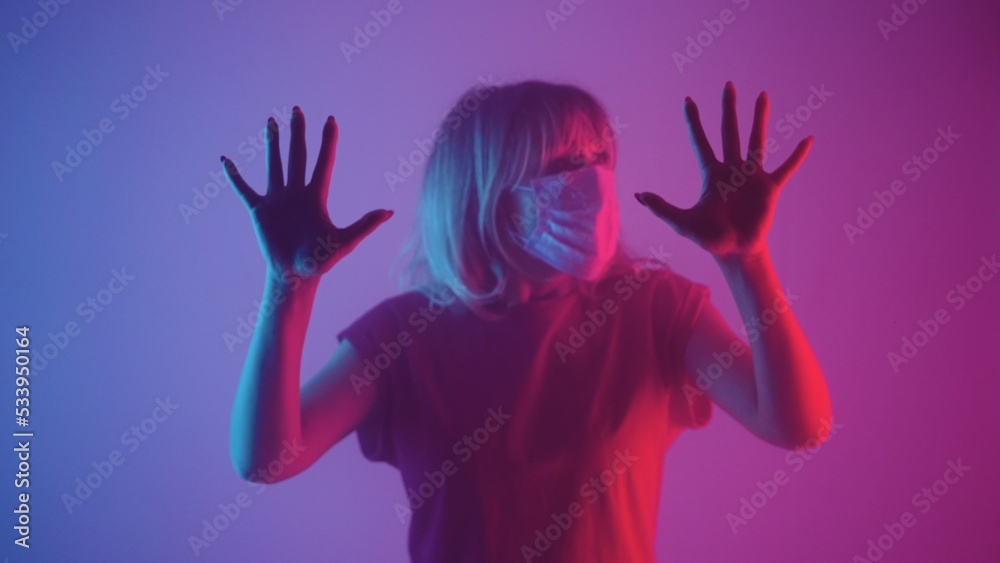Woman in medical mask looking to side and touching her palms on glass. An upset female touches glass with her hands in dark studio against backdrop of purple pink soft light and smoke. Close up. Woman in medical mask looking to side and touching her palms on glass. An upset female touches glass with her hands in dark studio against backdrop of purple pink soft light and smoke. Close up.
