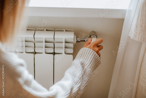 Young woman in long winter beige sweater is posing at home near the radiator