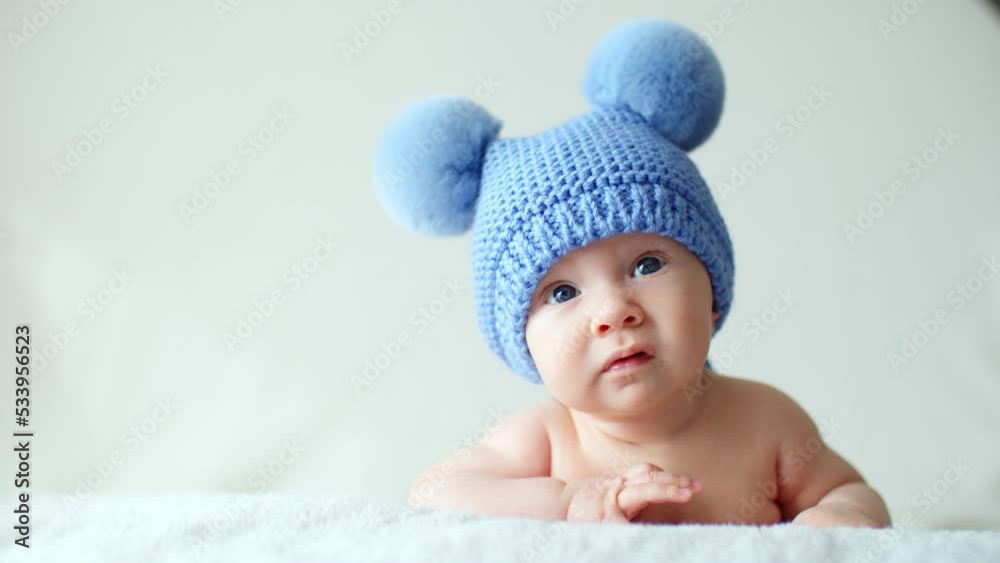 Calm two-month-old infant is lying on a blanket. Beautiful, cute little child with blue eyes looks into camera. Sweet newborn baby in a blue hat with pompoms looks into the camera. SLOW MOTION Calm two-month-old infant is lying on a blanket. Beautiful, cute little child with blue eyes looks into camera. Sweet newborn baby in a blue hat with pompoms looks into the camera. SLOW MOTION