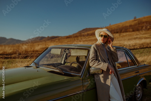 Elegant fashionable woman wearing trendy sunglasses, white hat, stylish autumn coat, posing outdoor, near green vintage car. Copy, empty space for text