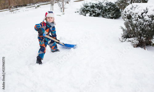 5-6 year old boy in bright warm overalls, helps to clear the yard with snow after a snowstorm, throwing snow away with a big shovel. Winter season, bad weather, cold, snowy