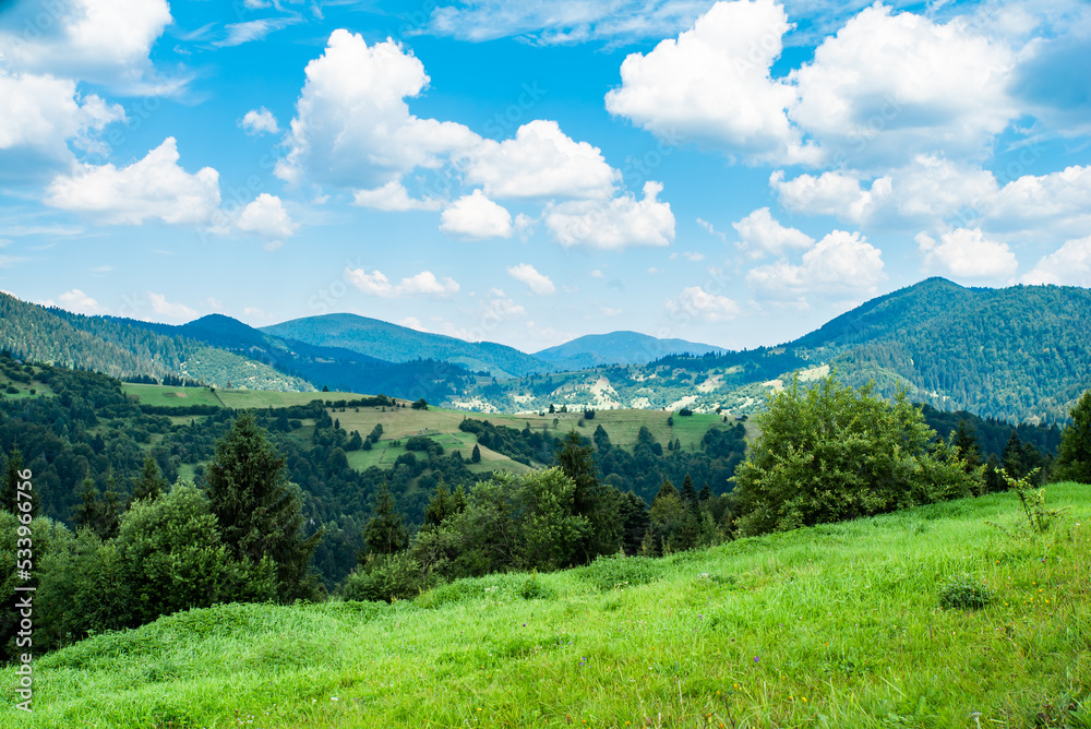 Scenic panoramic view of idyllic hills with blooming meadows, green fields on the hill against the background of mountain peaks on a beautiful sunny day with blue sky and clouds in spring