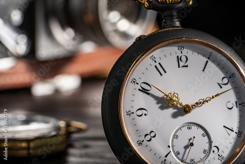 Silver antique pocket watch with gold hands and vintage film camera in background. An old round dial of a pocket watch on a dark wooden table and blurry camera lens with aperture. Close up.