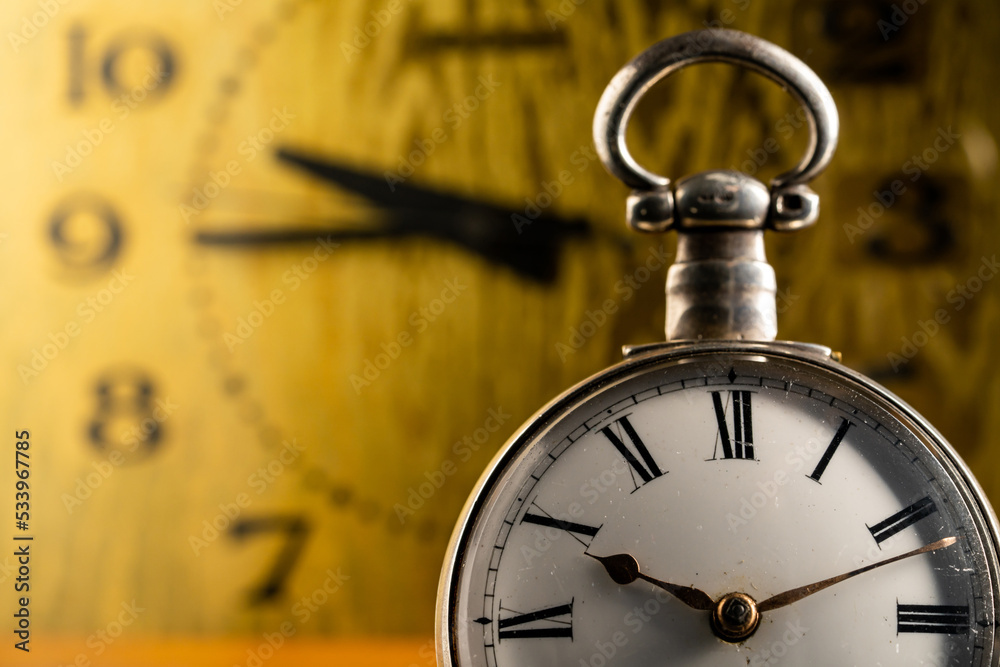 Silver antique pocket watch with Roman numerals and vintage table clock in blurry background. An old round white dial of pocket watch and yellow face of table clock with black hands. Close up. Silver antique pocket watch with Roman numerals and vintage table clock in blurry background. An old round white dial of pocket watch and yellow face of table clock with black hands. Close up.