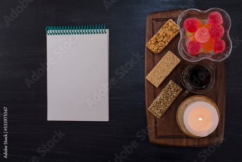 A glass of tea served with marmelades and sesame waffles on a wooden platter with a notebook aside