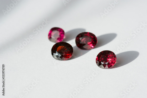 Genuine natural cherry red rhodolite garnets gemstones lot, set settings for making jewelry on bright daylight. Gemology mineralogy fashion theme. White paper background. Oval faceted. Semiprecious.
