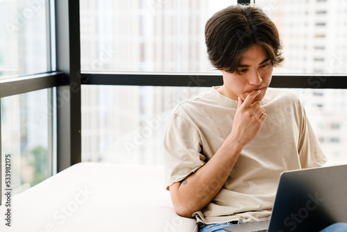 Asian young man using laptop computer while sitting by window at home