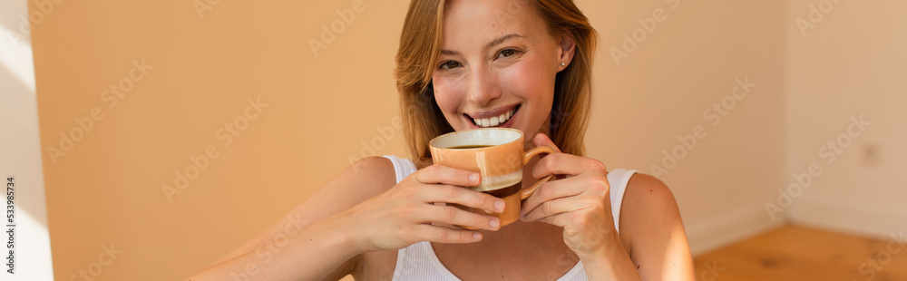 Positive blonde woman holding cup of coffee and looking at camera at home, banner. Positive blonde woman holding cup of coffee and looking at camera at home, banner.