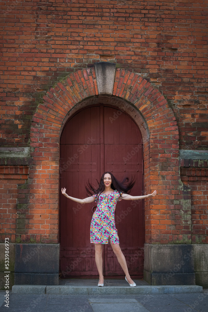 A beautiful slender smiling woman with long dark hair, in a light summer dress with a floral pattern, stands at the door in the form of an arch of an old red brick building.