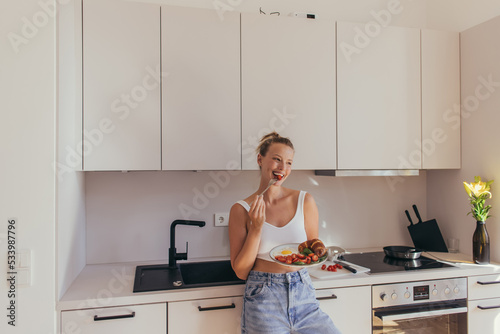Smiling blonde woman eating cherry tomato and holding plate with egg and croissant in kitchen.