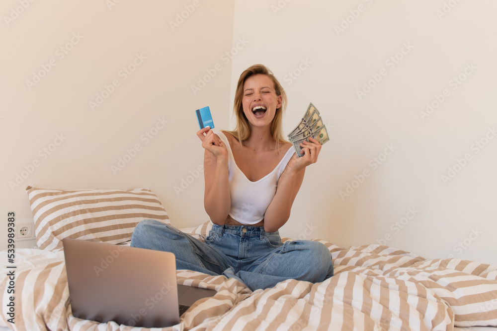 Excited blonde woman holding credit card and money near laptop on bed.