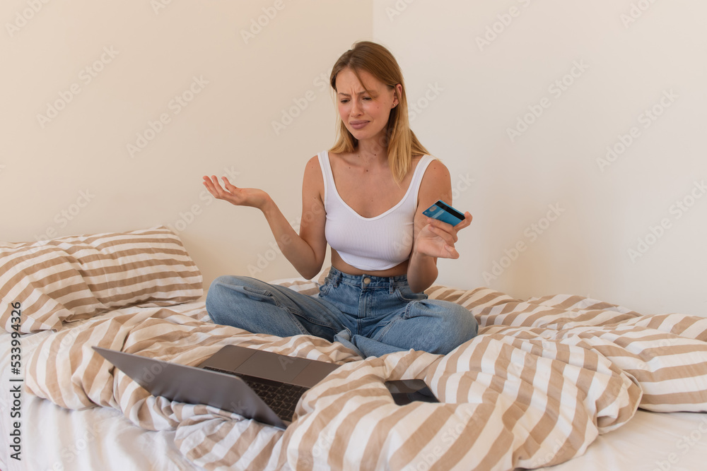 Upset woman holding credit card near laptop and smartphone on bed at home. Upset woman holding credit card near laptop and smartphone on bed at home.