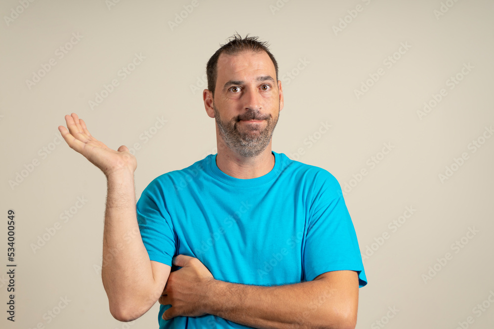 Hispanic man with a beard wearing a blue t-shirt in a skeptical attitude offering something in the palm of his hand, isolated over beige background. Hispanic man with a beard wearing a blue t-shirt in a skeptical attitude offering something in the palm of his hand, isolated over beige background.