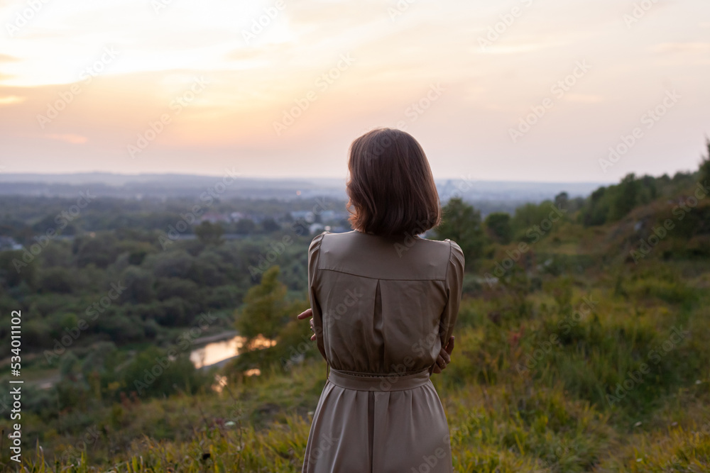 Beautiful young smiling girl in a long brown dress stands along the lawn. Happy woman walks at sunset on a hill overlooking the river. Concept of having rest in park during summer holidays or weekends