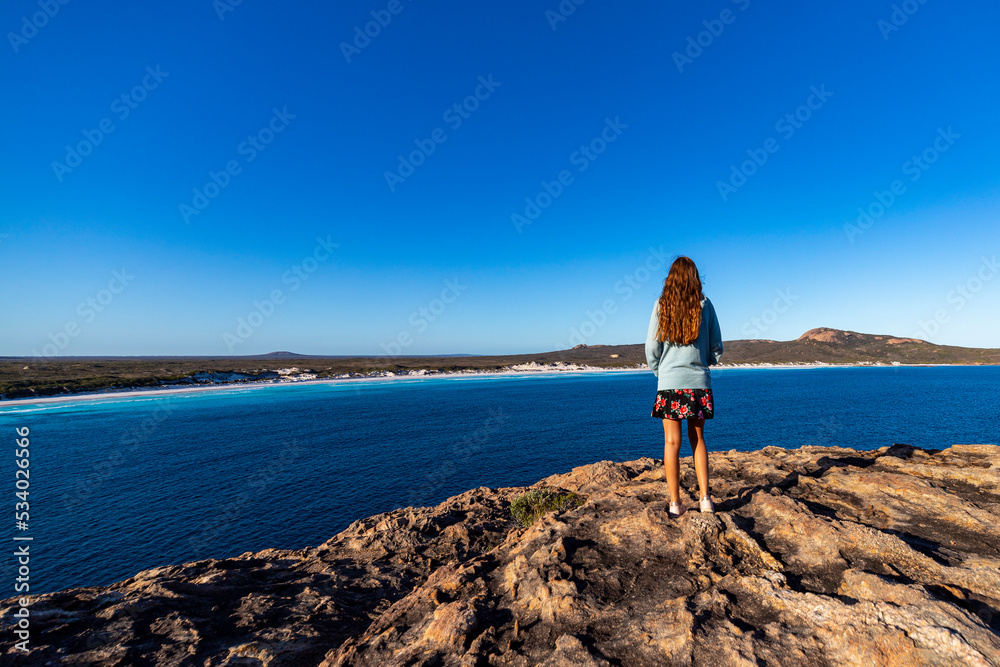 a long-haired girl in a dress stands at the top of a mountain admiring the view of a vast bay of white sand and turquoise water in cape le grand national park