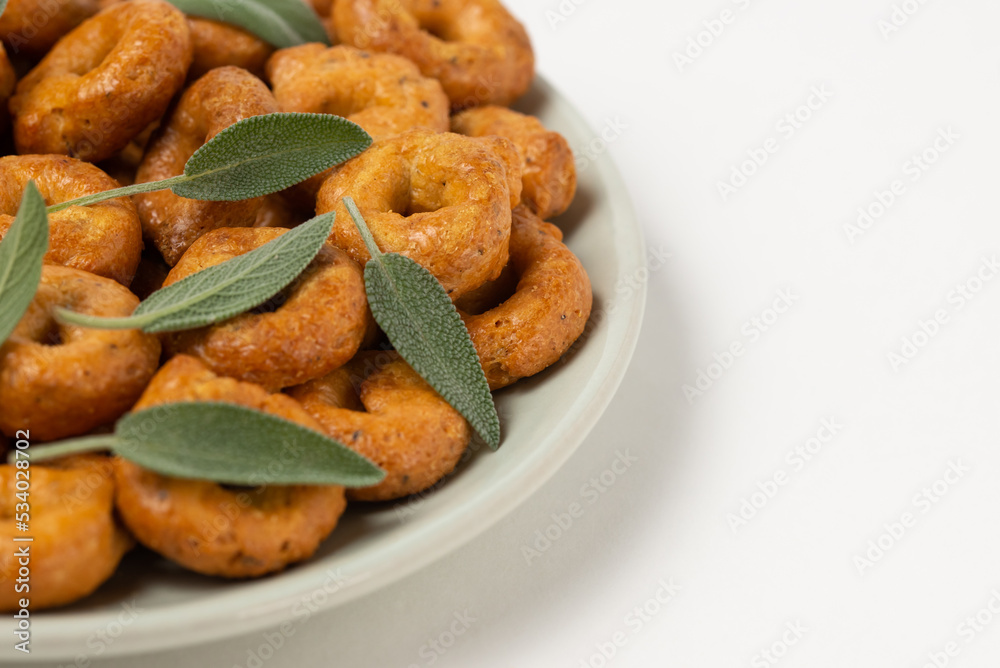 Traditional italian snacks taralli or tarallini with sage leaves in plate isolated on white background, close-up Traditional italian snacks taralli or tarallini with sage leaves in plate isolated on white background, close-up
