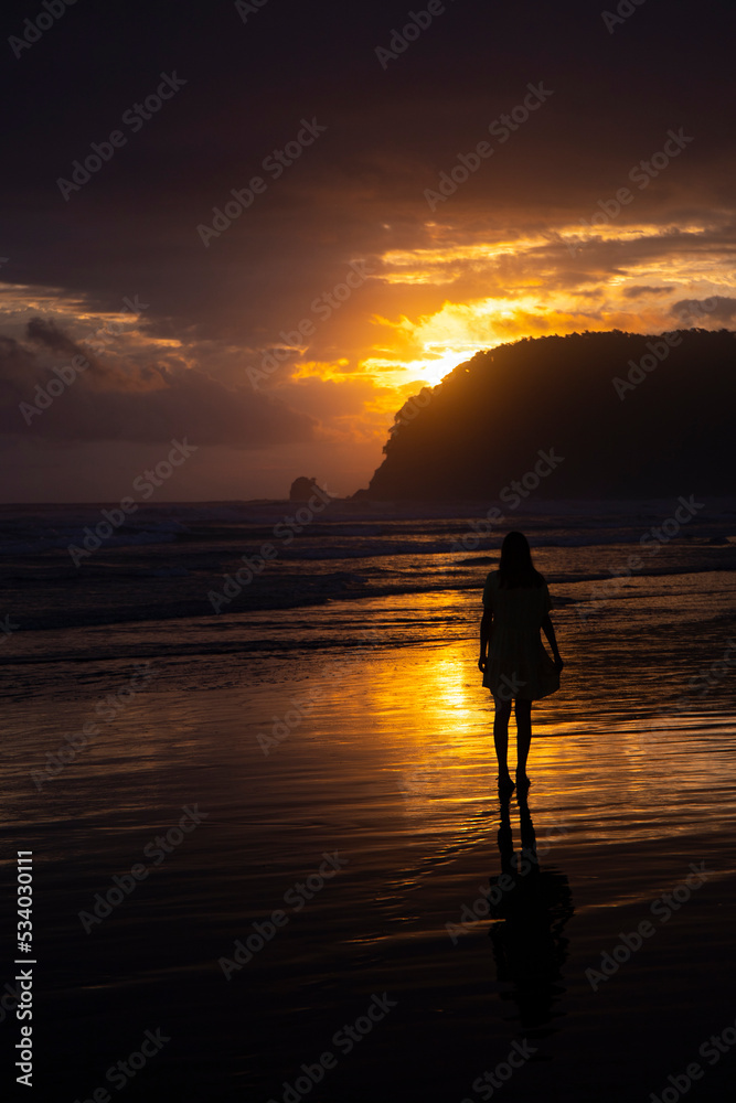 Fototapeta premium silhouette of a beautiful woman in a long dress with a colourful sunset on a tropical beach in the background; magical sunset over the ocean in costa rica, playa san miguel