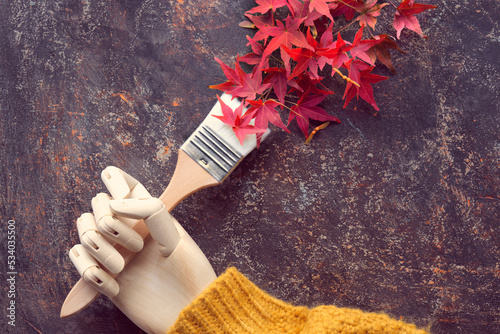 Paint brush, paintbrush with red oak leaves as paint. Brush in artistic model wooden hand in orange sweater. Autumntime colors, creative flat lay, top view.