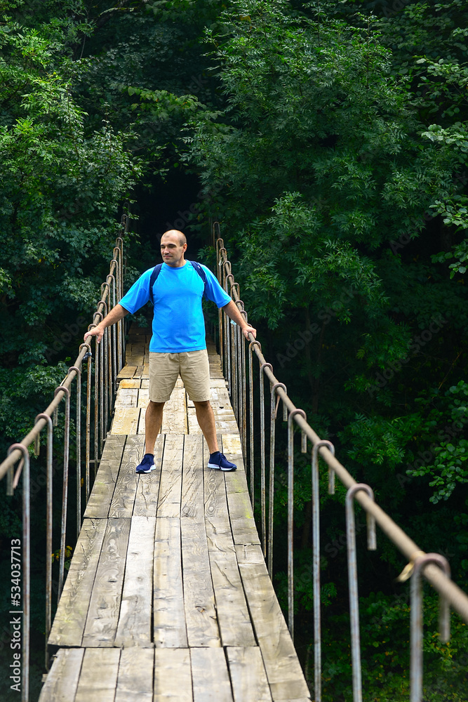 a male traveler with a backpack on his shoulders walks along a suspension bridge over a mountain river