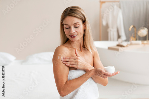 Body treatments. Happy middle aged woman applying moisturising cream on shoulder, making beauty care routine © Prostock-studio